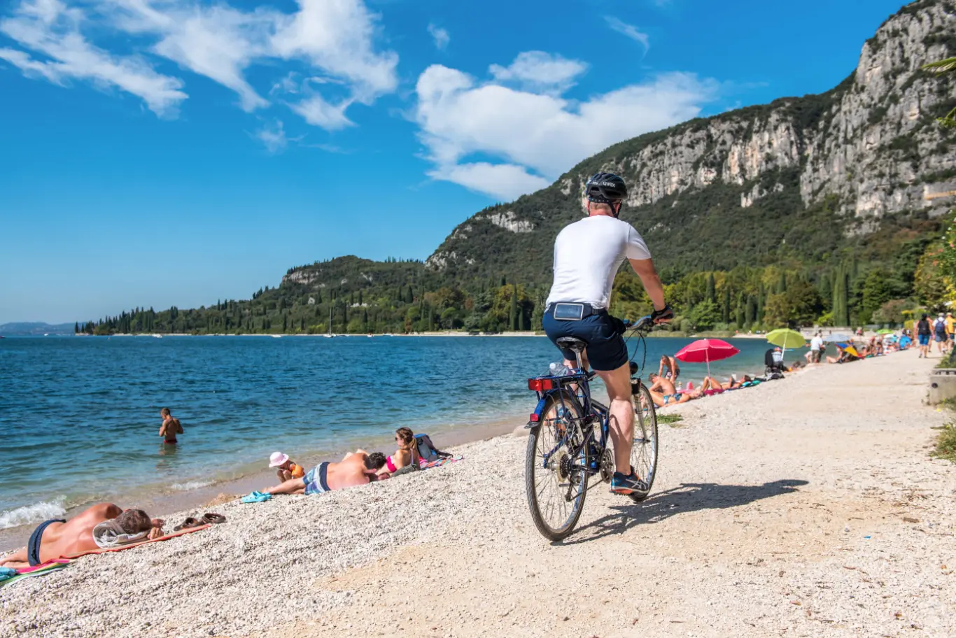 Ciclista su pista lungolago, bagnanti sulla spiaggia.